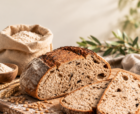 Une miche de pain complet rustique faite maison, partiellement tranchée pour révéler une mie tendre et alvéolée, repose sur une planche à découper en bois, aux côtés de quelques grains éparpillés et d'un sac de farine en toile de jute.